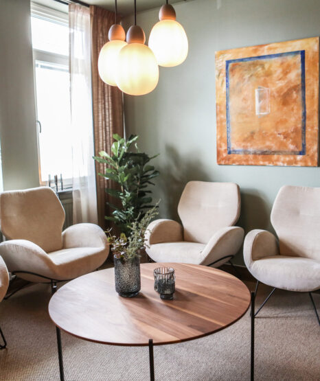 Ett modernt vardagsrum med fyra beige stolar, ett runt soffbord i trä, växter, ett stort fönster, väggmålning och tre klotlampor är det perfekta utrymmet för avkoppling. Inspirerad av den lugna atmosfären på tandläkare Övik privata kontor.