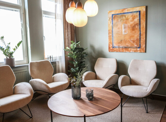 Ett mysigt vardagsrum med moderna möbler, inklusive fyra beiga fåtöljer, ett runt soffbord i trä, tre hänglampor, en väggmålning och krukväxter – perfekt för att koppla av efter ditt besök hos tandläkare örnsköldsvik.